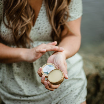 A woman with long brown hair and a green polka-dot dress sits beside the ocean and applies an open tin of Calm Body Balm to the inside of her wrist